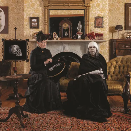 A staged Victorian-style interior with patterned wallpaper and antique furniture, where two women (artists megan evans and Maree Clarke) dressed in black sit together on an upholstered chair.