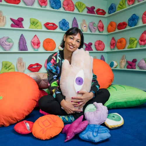 Artist Kate Beynon sits on a blue carpet in front of light teal shelf with colourful soft sculptures of body parts and other motifs. Some have Chinese characters written on them. Kate sits amongst larger soft sculptures and holds one in her arms, which appears to be the back of a hand with an eye on it. Kate is smiling at the camera. 