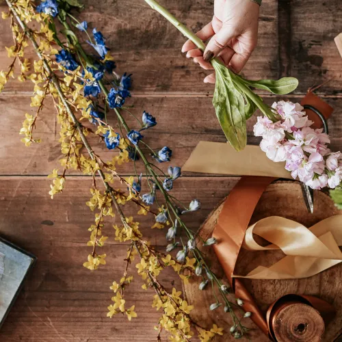 A wooden table scattered with floral arrangement materials, including long stems of yellow and blue flowers, pink blossoms held by a hand, and green leaves. Surrounding the flowers are ribbons in shades of brown and beige, a round wooden board, a small mirror and a box with twine. 