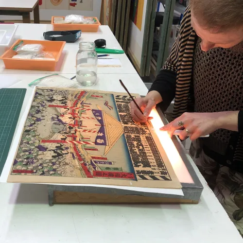 A conservator patches up a torn edge of a yellowed Japanese print using a small paint brush. The print is placed on top of a lightbox. 