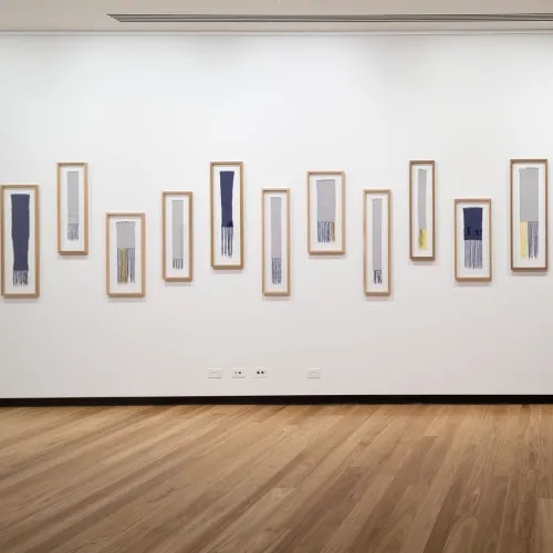 A photograph of an installation of framed small tapestries on a white wall.