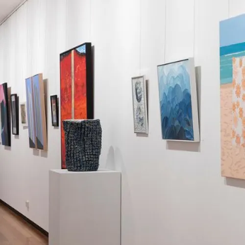 The image shows a long, well-lit gallery space with wooden flooring and white walls adorned with various artworks. The artworks include colourful abstract paintings, geometric compositions, and landscape scenes, all framed or mounted on the walls. The perspective extends down the hallway.