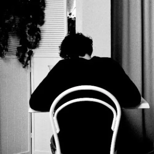 A black-and-white photo of a person seated alone at a table in a cafe. They are facing away from the camera. The space is filled with white chairs and tables, and an arched curtain feature dominates the background. Indoor plants add contrast to the scene.