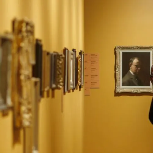 A warmly lit gallery corridor with golden-yellow walls and wooden floors. A long row of small, framed artworks are on the wall to the left, shown in soft focus. At the far end of the space, more framed portraits and small works hang on the wall. Several visitors walk through the gallery, some pausing to look closely at the artwork, while others continue moving. 