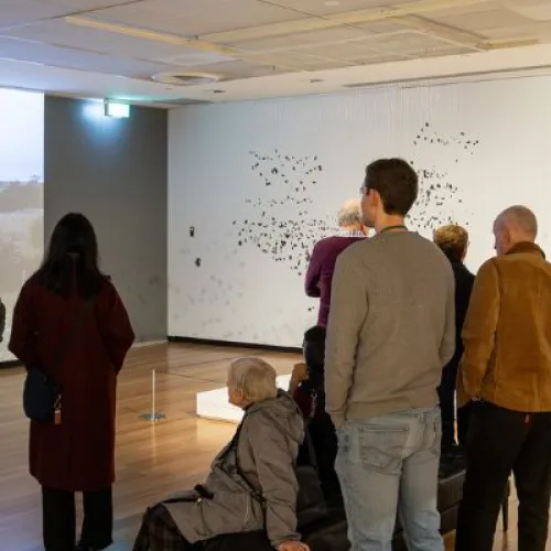 A group of people stand and sit throughout a large gallery with white walls and wooden floors. On the left wall, a large projected video shows an outdoor landscape with a burning object. In the centre, visitors are standing facing the projection and a white wall displaying a delicate installation made of many small black elements suspended in a scattered pattern. Additional framed works hang on the right wall. 
