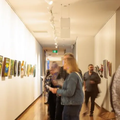 A long gallery corridor with wooden floors and rows of colourful artworks hung on both walls. Several people move through the space, some appearing blurred from motion, while others pause to look at the paintings under bright track lighting.
