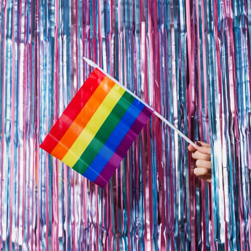 A rainbow flag is being held in front of a background of purple and pink streamers