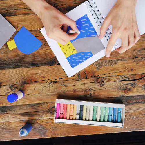 A selection of pencils, crayons, scissors and paper sit on a wooden table with 2 hands visible, 1 holding a crayon