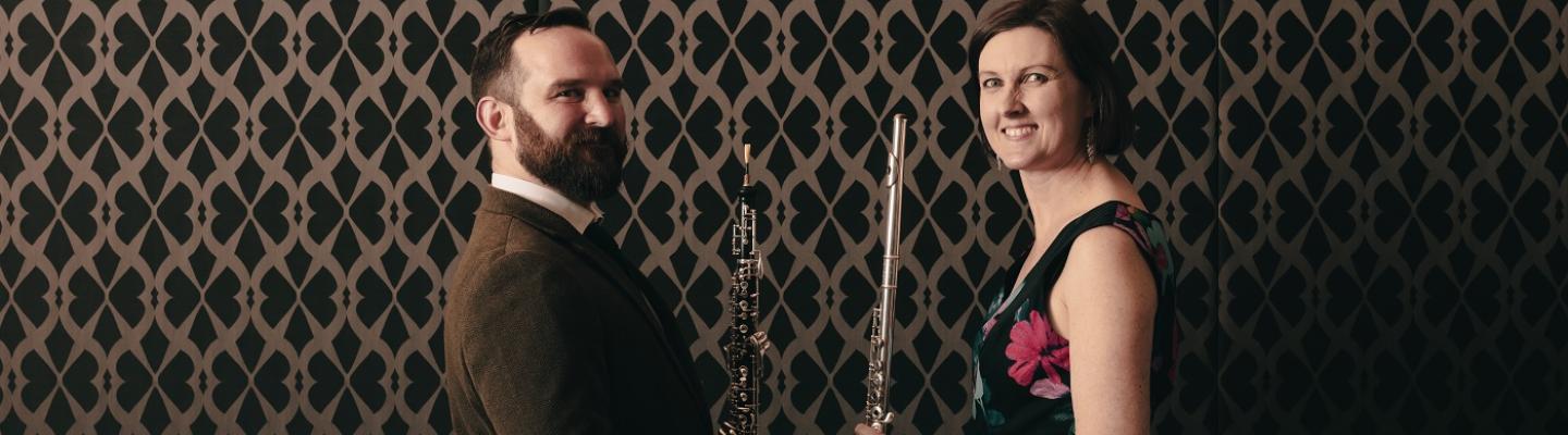 Two musicians posing for the camera, with a patterned wallpaper behind them