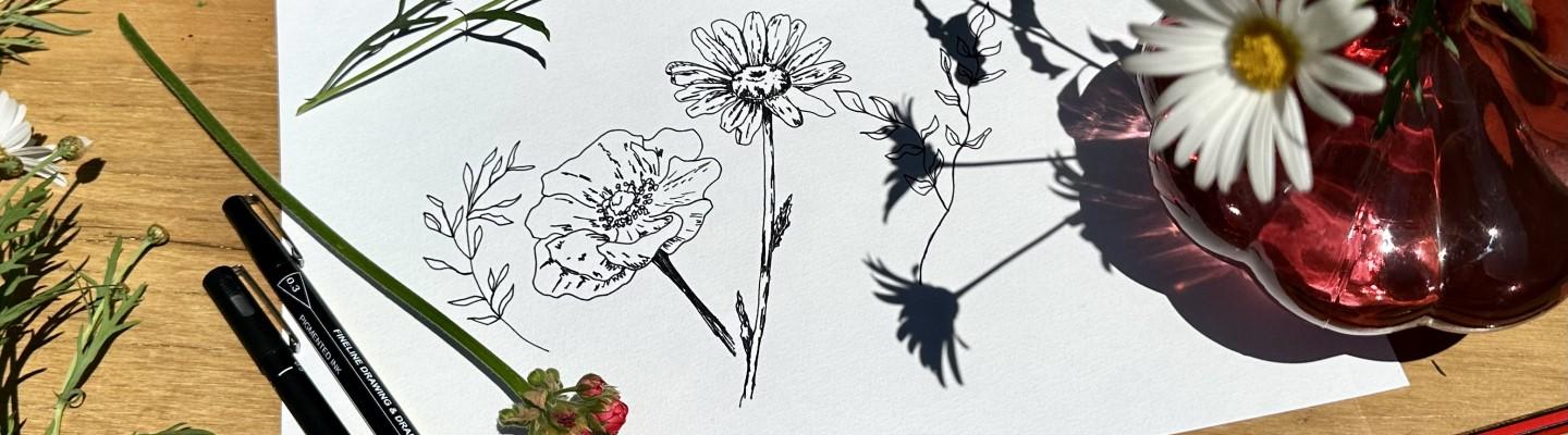 Ink drawing of flowers on white piece of paper, next to pens and real flowers.