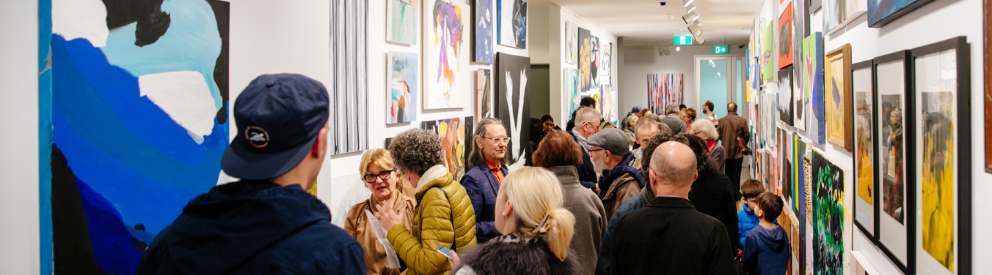 A hallway filled with a large crowd of people talking and looking at the artwork which covers both walls