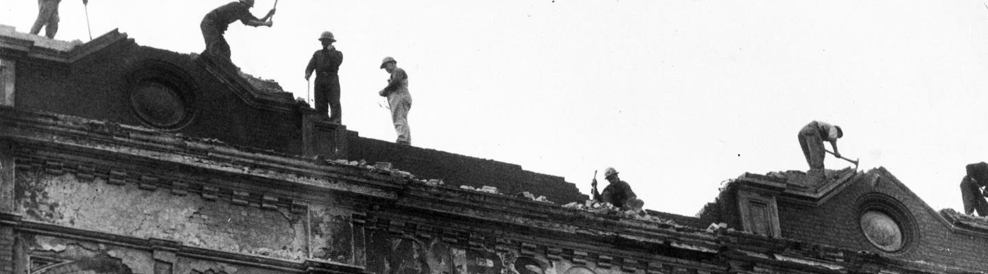 A black and white photo of people working on top of an old building facade which is damaged and burnt