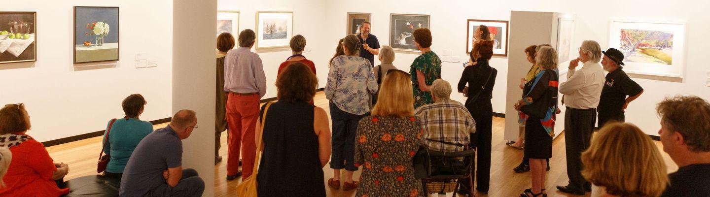 A group of people standing in an art gallery space with art handing on the walls, while listening to a talk