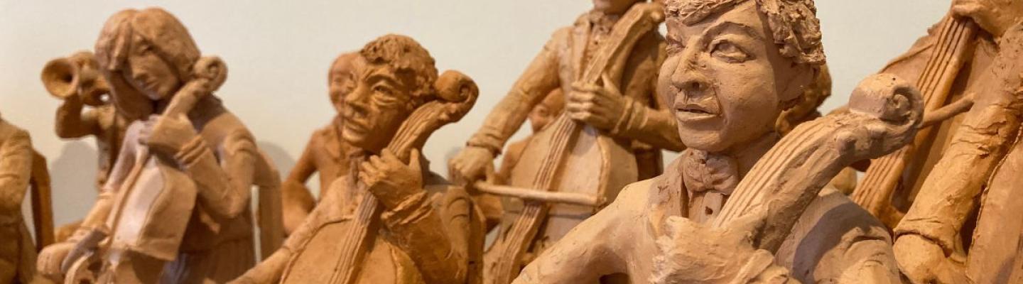 Unpainted clay sculptures of five figures playing cellos. Three of the figures are seated while the remaining two are standing up, behind them. In the background is another clay figure, standing and blowing a horn and another seated figure whose instrument is obstructed from view. 