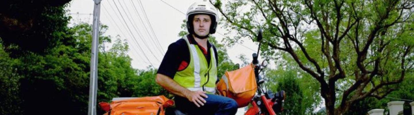 A postal worker wearing a yellow hi-vis vest and white helmet, and riding a red motorcycle with orange postal carrier bags on the side
