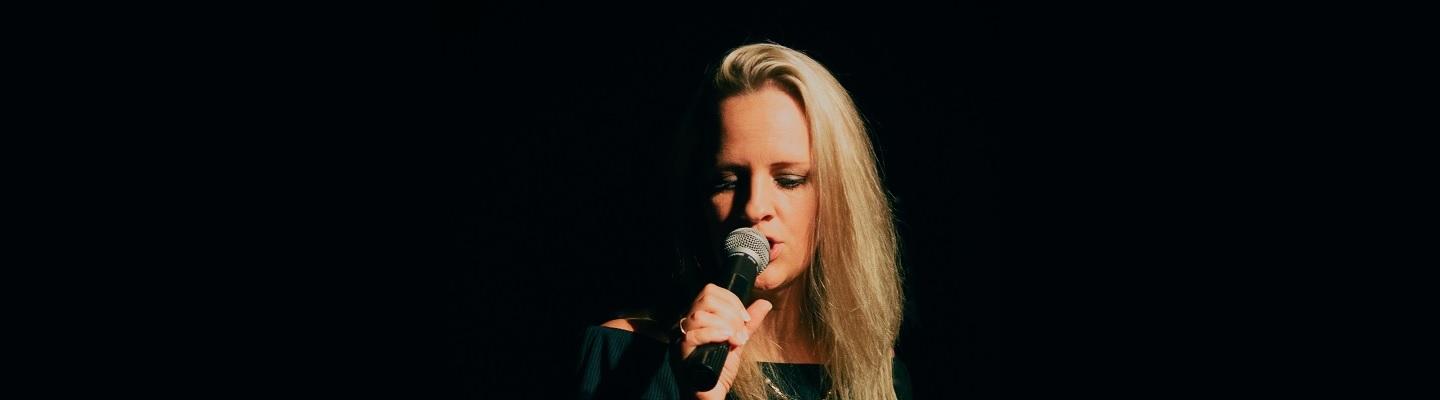 half of the profile of a blonde woman singing into a microphone against a black background