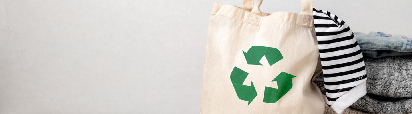 A canvas tote bag with a recycling symbol on the front and with a sleeve of clothing coming out the top of the bag