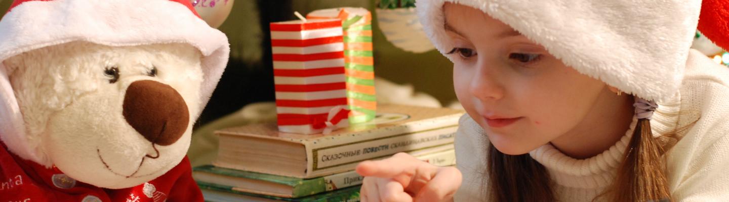 Young girl with Santa hat on is reading an opened book to a teddy placed next to her. Background with books and candle in front of a christmas tree