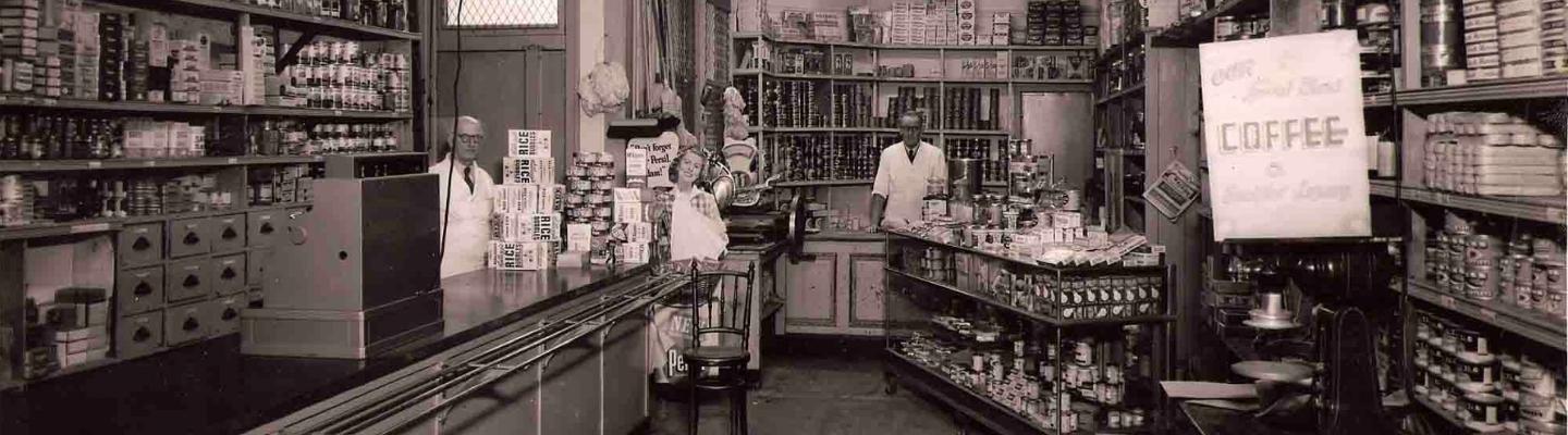 A sepa photograph of an old milk bar. Items can be seen on the shelves. 2 people are standing in the back wearing white