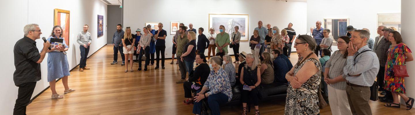 a crowd of people in a gallery space listen to a person speaking with a microphone in front of a wall of paintings