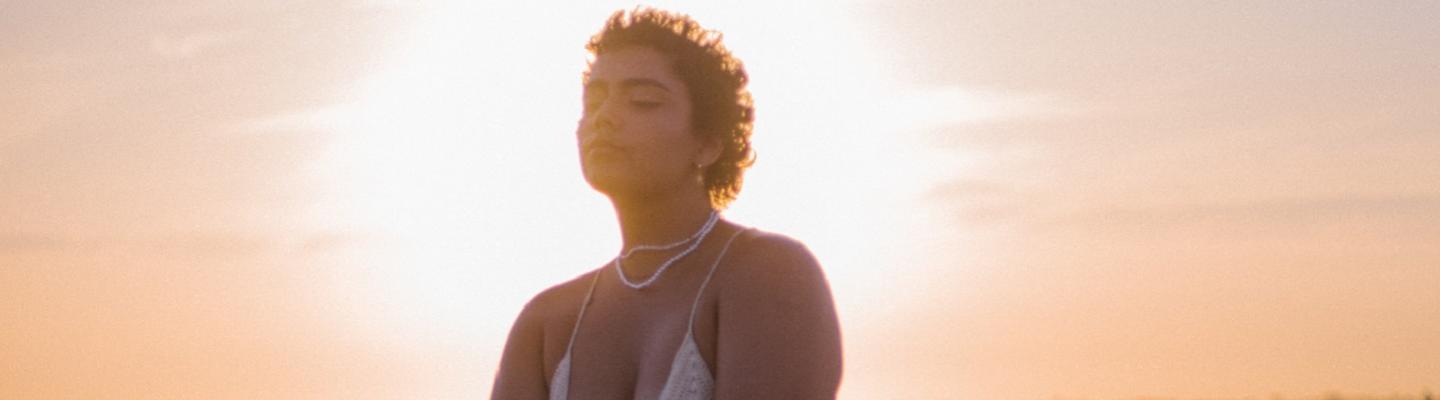 a person in a halter top and beaded necklace against a setting sun 