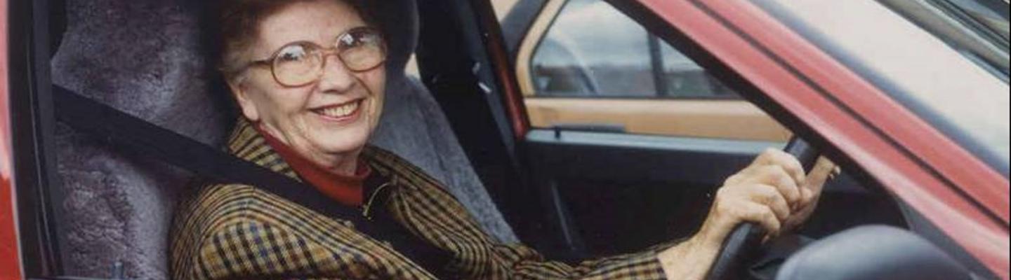 An older woman in a tweed jacket smiles in the driver's seat of a red car