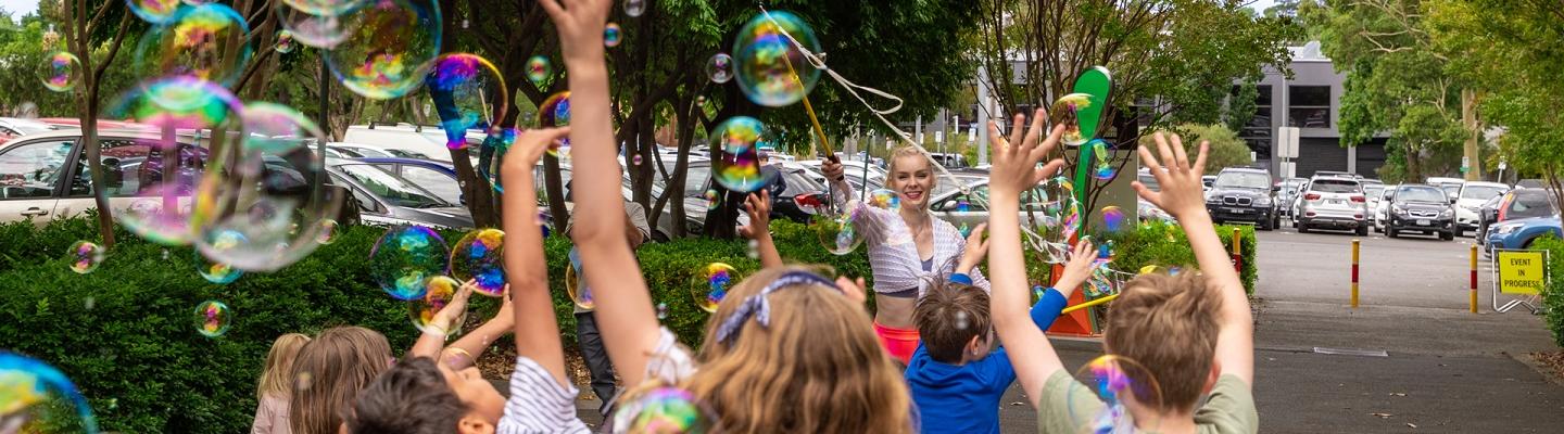 a group of kids jump to catch large, colourful bubbles being blown by a performer with a bubble want