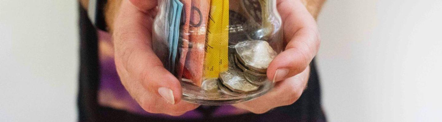 A close up of a pair of hands holding a glass jar of cash and coins