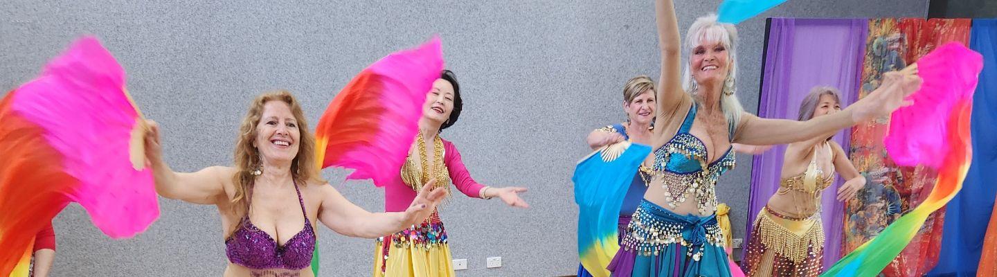 Five ;adies dancing. They are wearing belly dance constume and waving large, colourful flags