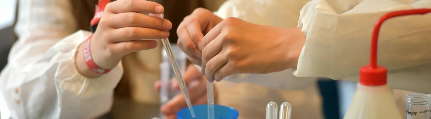 Close up view of 2 children holding pipette's in a blue cup with other science experiment items nearby.