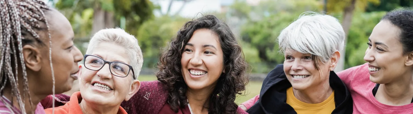 Five women are standing outside and smiling. The women are all wearing casual clothes and are standing with their arms around each other. They are all different ages and ethnicities.