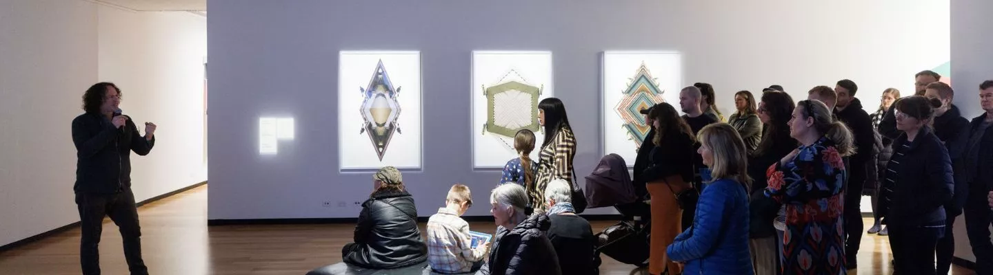 Image of man in gallery speaking to a crowd of people. Artwork is hung on wall in background. 