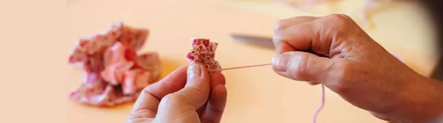 Photo of hands tying a pink piece of yarn around a small bunched up piece of pink patterned fabric. A blurry fabric flower is positioned in the background behind the hands.