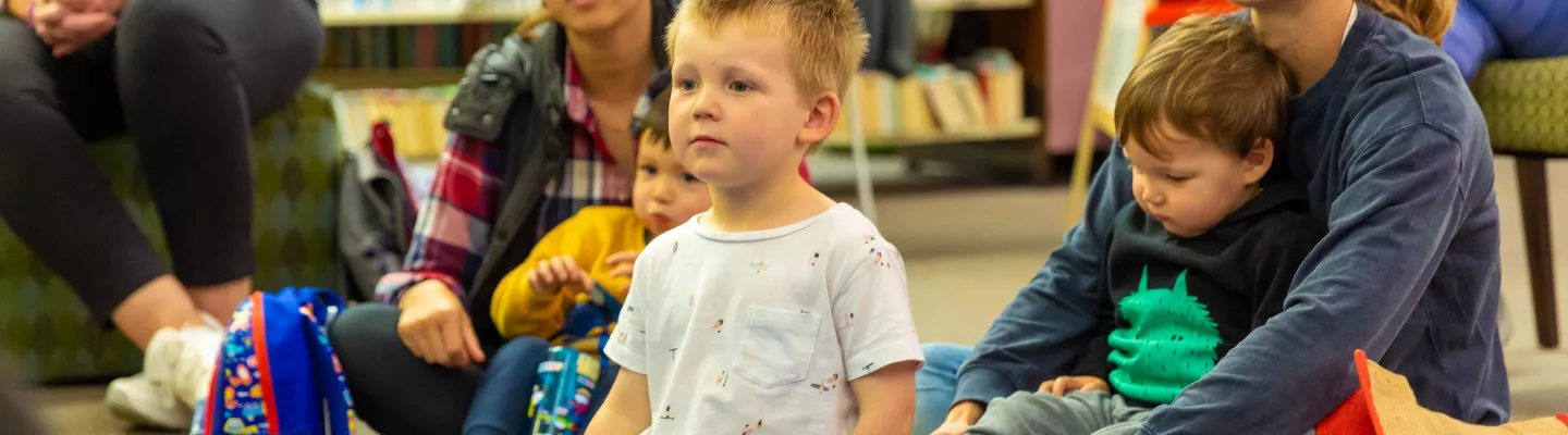 A group of children and their carers sit on a blue lap in a library.