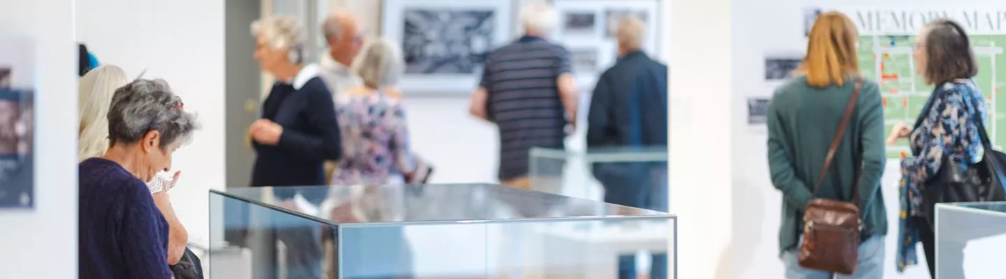 This image depicts a group of figure slooking through a gallery exhibition. The exhibition includes walls with pictures and print and a glass cabinet with historical memorbilia displayed