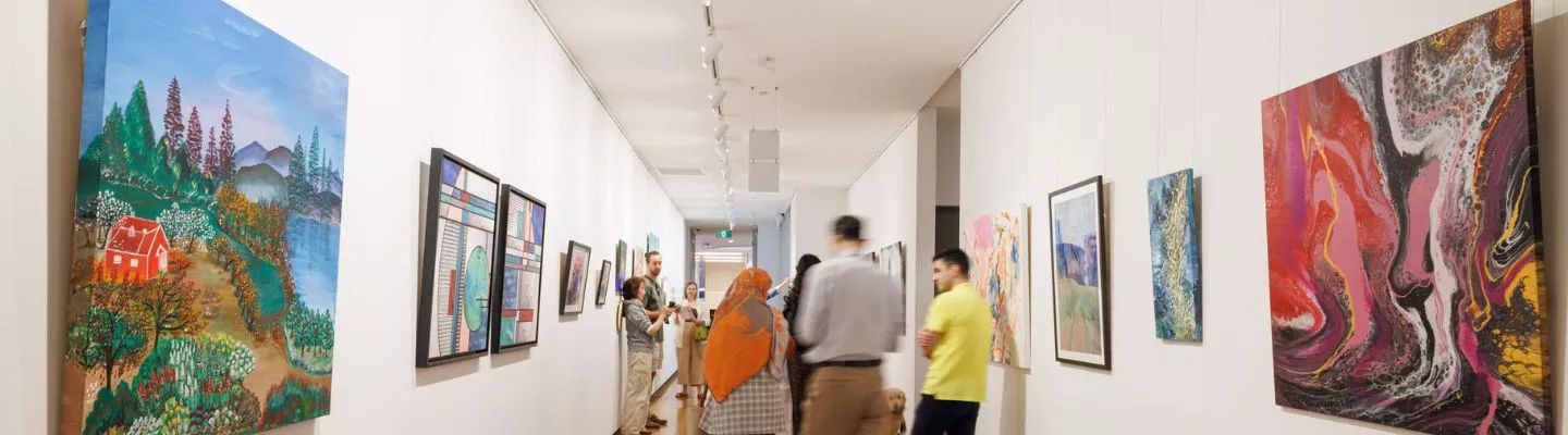 The image shows a long, well-lit gallery space with wooden flooring and white walls adorned with various artworks. The artworks include colourful abstract paintings, geometric compositions, and landscape scenes, all framed or mounted on the walls. Visitors are walking through the space, some engaged in conversation while others observe the pieces. Track lighting runs along the ceiling, illuminating the artworks. The perspective extends down the hallway.