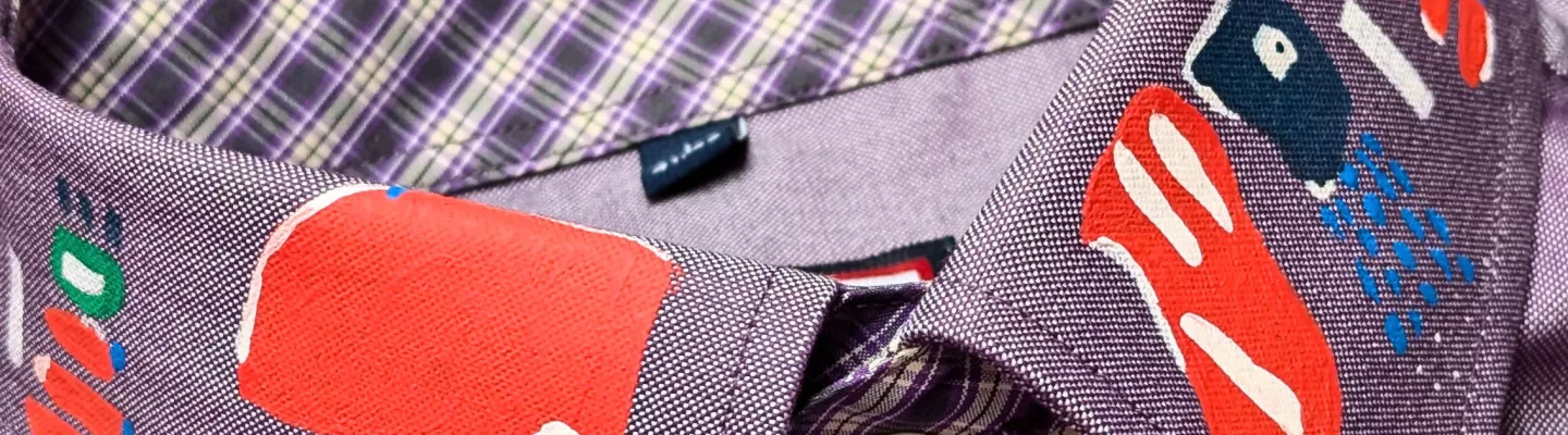 Close-up photograph of a buttoned-up purple shirt. The collar has been embellished with red, yellow, blue, green and white graphic shapes and patterns. 