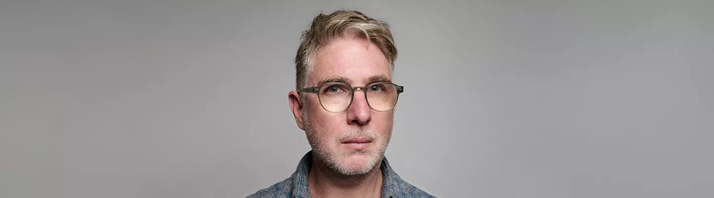 A portrait of David Rosetzky, a middle-aged man with short, light grey hair and glasses, wearing a checkered blue shirt over a grey t-shirt. He is looking directly at the camera with a neutral expression.
