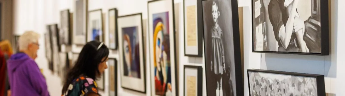 image of a gallery with assorted paintings hanging on the walls. Gallery visitors look closely at the paintings. 