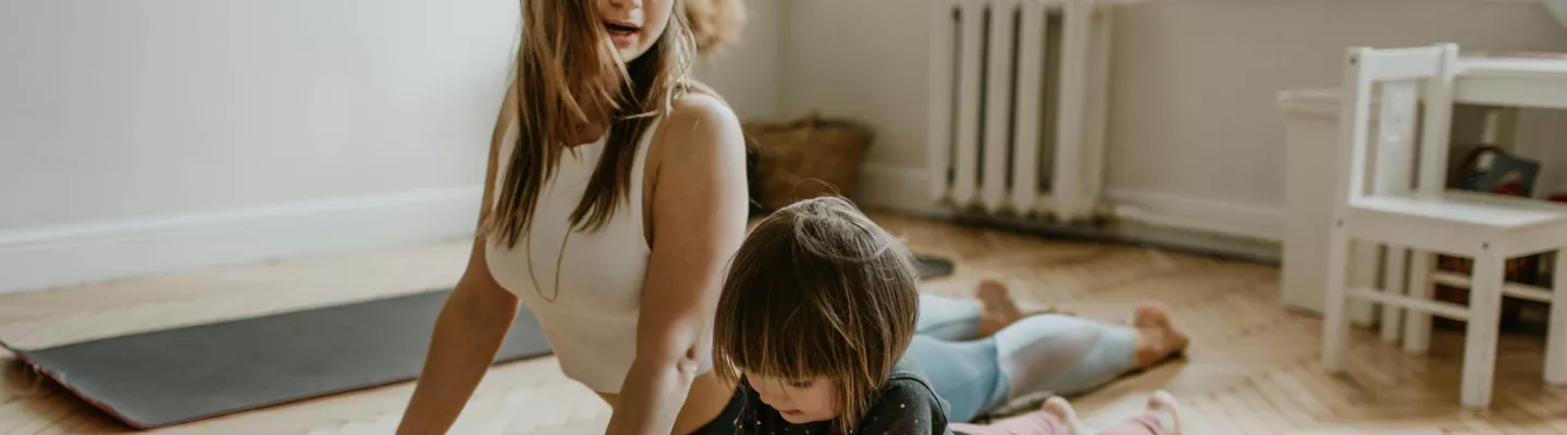 A woman and child are stretching on the floor, stomach to the floor with their legs outstretched and their head and chest lifted from the ground.
