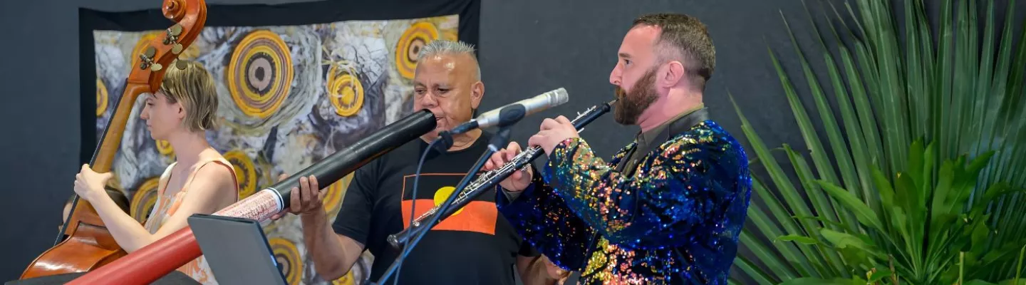 Three people performing. A man in a sparkly blue jacket plays the flute. Another man in a black t-shirt featuring the Aboriginal flag is playing the didgeridoo and a woman in the background plays cello