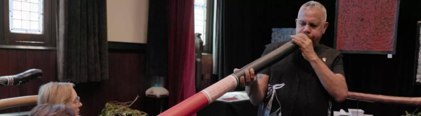 A man is standing playing a didgeridoo which is pointed at a seated woman's chest