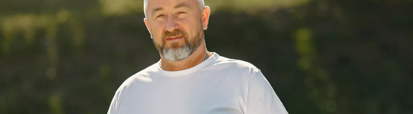 A man is wearing a white tshirt holding a drink bottle in one hand and a yoga mat tucked under the other arm.