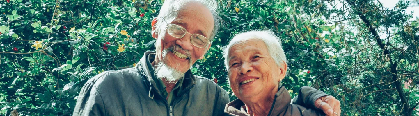 An elderly couple are smiling at the camera with one person's arm resting on the shoulder of the other person.