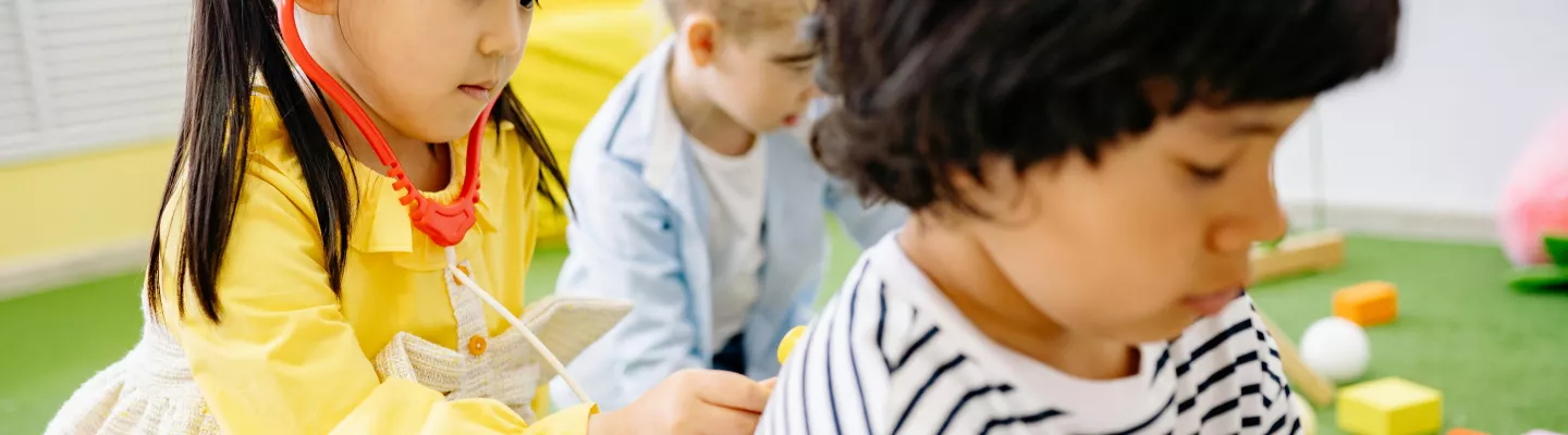 Girl in yellow dress playing doctor by pressing stethoscope onto boy's back.