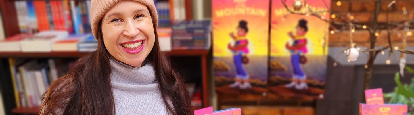 A smiling lady wearing a pink beanie and lilac jumper is holding a book in one hand and a take away coffee cup in another.
