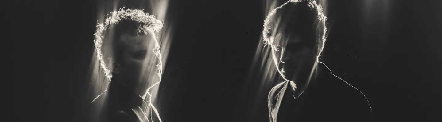 2 people standing in darkness, a hazy light shining from behind them illuminating the sides of the their faces and heads