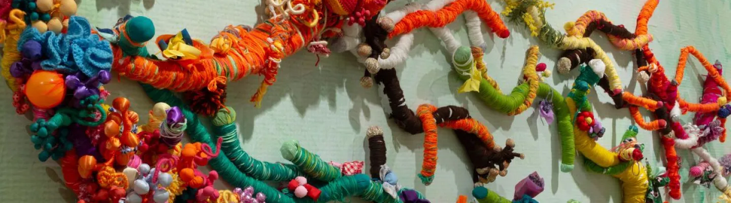 image of scultpural textile work on wall. The work is made up of varied bright colours of wool, mainly orange, gree, yellow, brown and pinks hues. They appear like vines on the wall. 