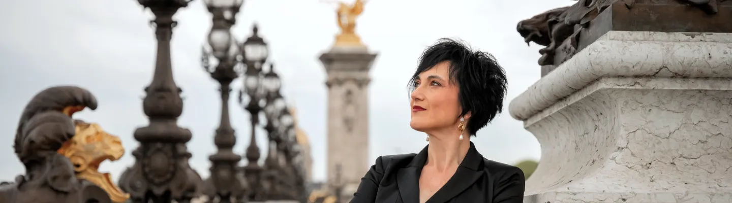 woman wearing black with short hair, standing on a bridge with ornate lamp posts.