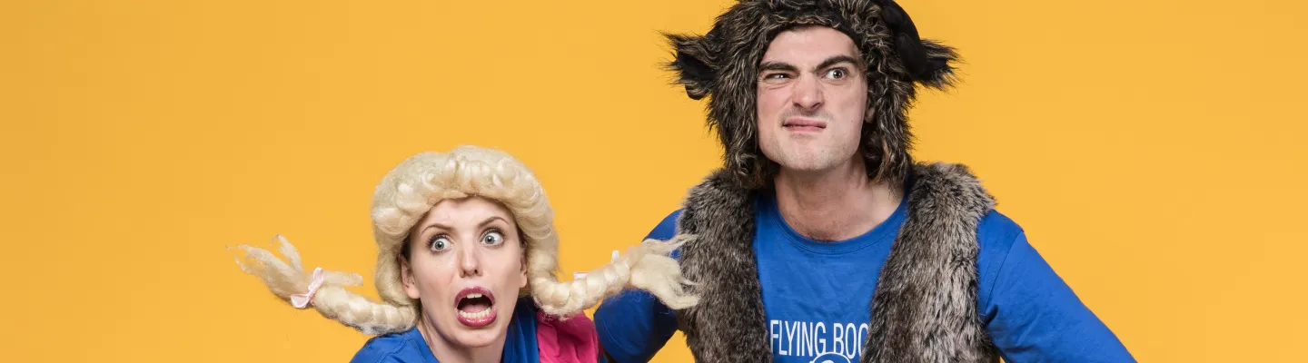 Two adults in costume. One is wearing a blond, plaited wig with a pink dress and gingham apron. The other has a furry headpiece and vest over a blue tshirt. Both are making funny faces.